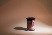 The Bridgewater Candle Company Hello Night 8oz Jar Candle features pinkish soy-blend wax in a small jar with a black lid and hanging tag, softly glowing on a plain surface against a neutral background with subtle curved light patterns.