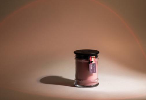 The Bridgewater Candle Company Hello Night 8oz Jar Candle features pinkish soy-blend wax in a small jar with a black lid and hanging tag, softly glowing on a plain surface against a neutral background with subtle curved light patterns.