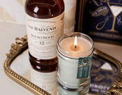 An Amber & Emerald 8oz Jar Candle by Bridgewater Candle Company sits lit on a tray beside a bottle of The Balvenie DoubleWood 12-year-old Scotch; a green tag hangs from the candle and part of a cocktail-themed book is visible behind.