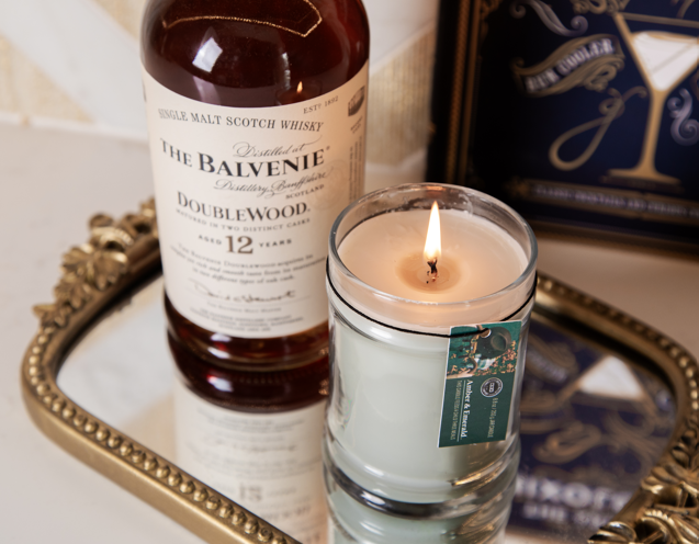 An Amber & Emerald 8oz Jar Candle by Bridgewater Candle Company sits lit on a tray beside a bottle of The Balvenie DoubleWood 12-year-old Scotch; a green tag hangs from the candle and part of a cocktail-themed book is visible behind.