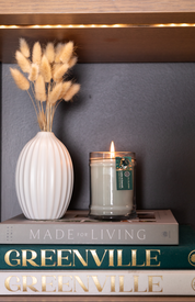 A white ribbed vase with dried grass, a lit Amber & Emerald 8oz Jar Candle by Bridgewater Candle Company in a glass jar, and two books—“MADE FOR LIVING” on top and “GREENVILLE” below—are stacked on a shelf.