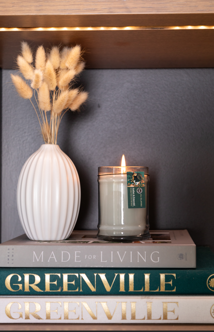 A white ribbed vase with dried grass, a lit Amber & Emerald 8oz Jar Candle by Bridgewater Candle Company in a glass jar, and two books—“MADE FOR LIVING” on top and “GREENVILLE” below—are stacked on a shelf.
