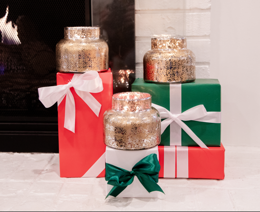 Three gold candle holders rest on red and green ribbon-wrapped gifts before a glowing fireplace, creating a festive scene perfect for the Christmas Bliss Holiday Mercury Jar Candle by Bridgewater Candle Company, featuring soy-blend wax.