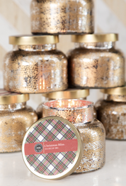 A glowing Christmas Bliss Holiday Mercury Jar Candle by Bridgewater Candle Company, in gold-speckled glass and soy-blend wax, sits before a stack of similar unopened jars with festive plaid labels.