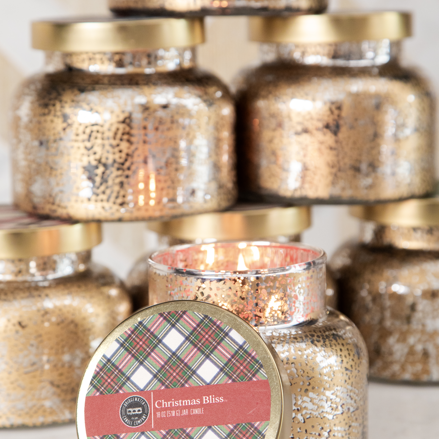 A glowing Christmas Bliss Holiday Mercury Jar Candle by Bridgewater Candle Company, in gold-speckled glass and soy-blend wax, sits before a stack of similar unopened jars with festive plaid labels.