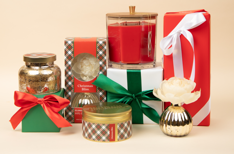 A festive holiday display features the Bridgewater Candle Company Christmas Bliss Holiday Large Decorative Candle, wrapped gifts with red, green, and white ribbons, a red glass jar candle, floral diffuser, and gold-accented jars on beige.