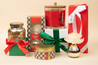 A festive holiday display features the Bridgewater Candle Company Christmas Bliss Holiday Large Decorative Candle, wrapped gifts with red, green, and white ribbons, a red glass jar candle, floral diffuser, and gold-accented jars on beige.