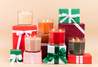 The Christmas Bliss Holiday Large Decorative Candle by Bridgewater Candle Company glows in a glass holder, surrounded by wrapped gift boxes with green, red, and white ribbons on a beige background.