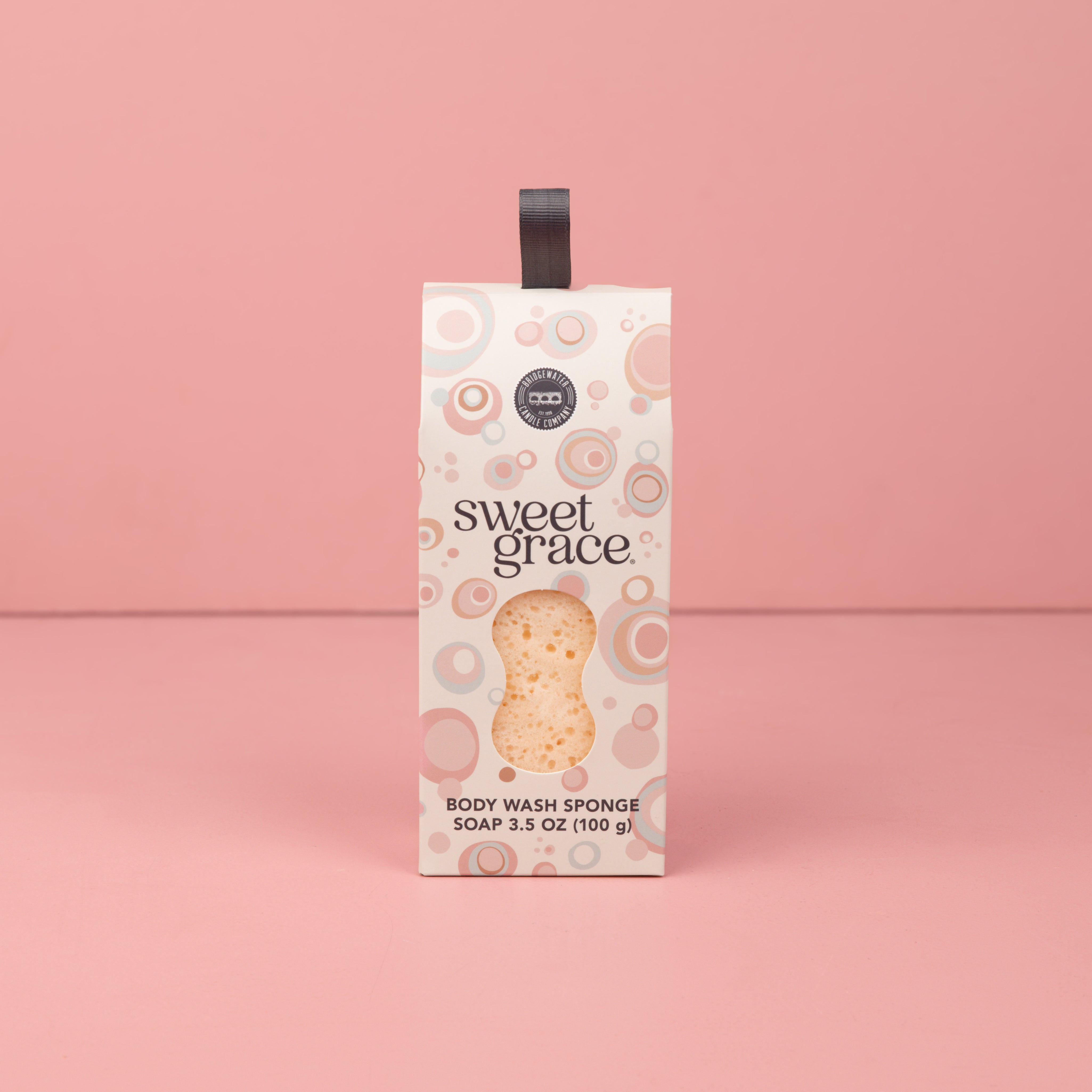 The Bridgewater Candle Sweet Grace Body Wash Sponge stands upright against a plain pink background, its patterned light packaging with a dark handle offering creamy lather for gentle skin exfoliation.