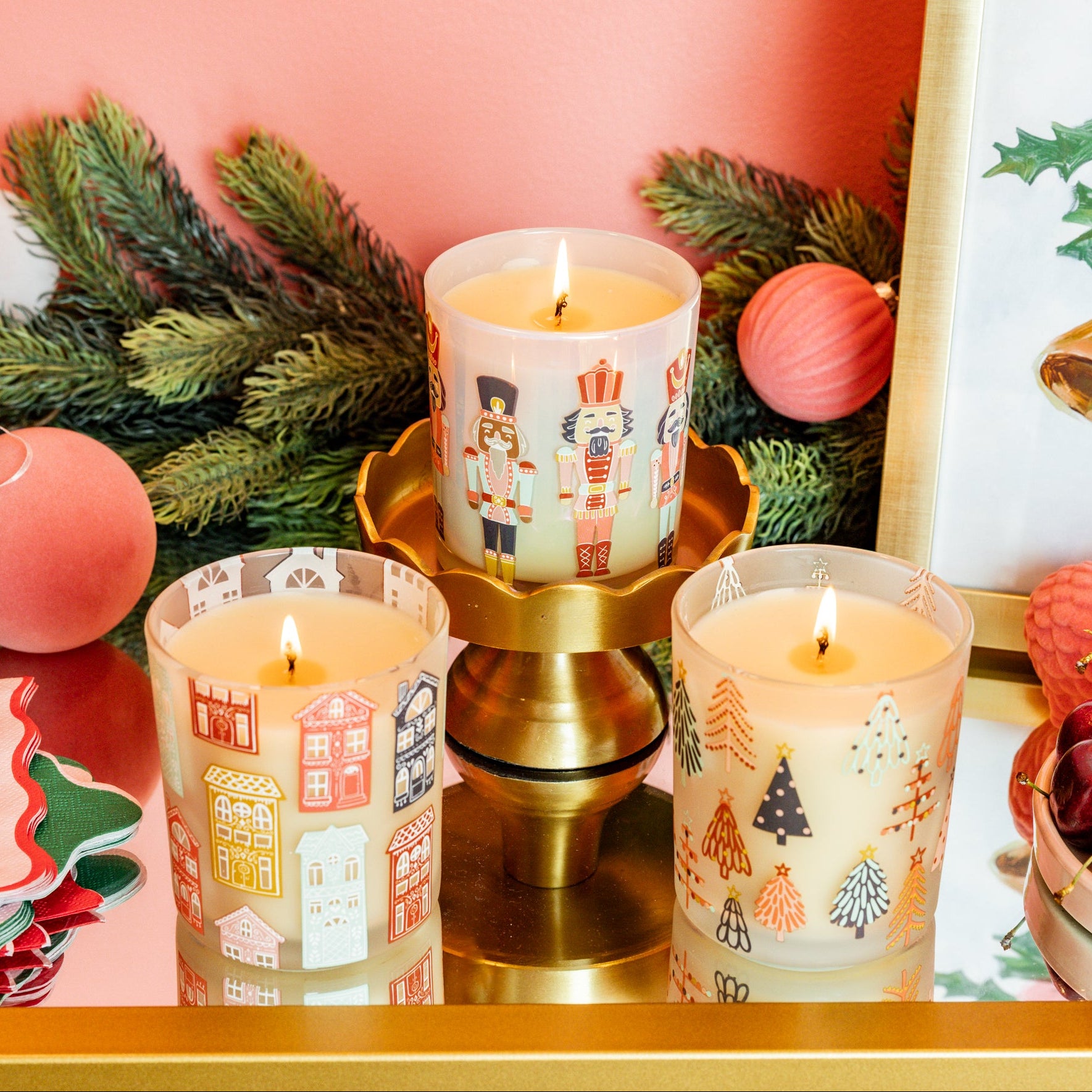 Three festive holiday candles, including the Bridgewater Candle Sweet Grace Holiday Nutcracker Candle made from soy-blend wax, are lit on a gold stand amid pine branches, red ornaments, a framed bell print, and cherries atop a mirrored surface.