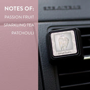 A Sweet Grace Auto Vent Clip by Bridgewater Candle Company, featuring angel wings, fills your car with passion fruit, sparkling tea, and patchouli fragrance notes against a pink backdrop.
