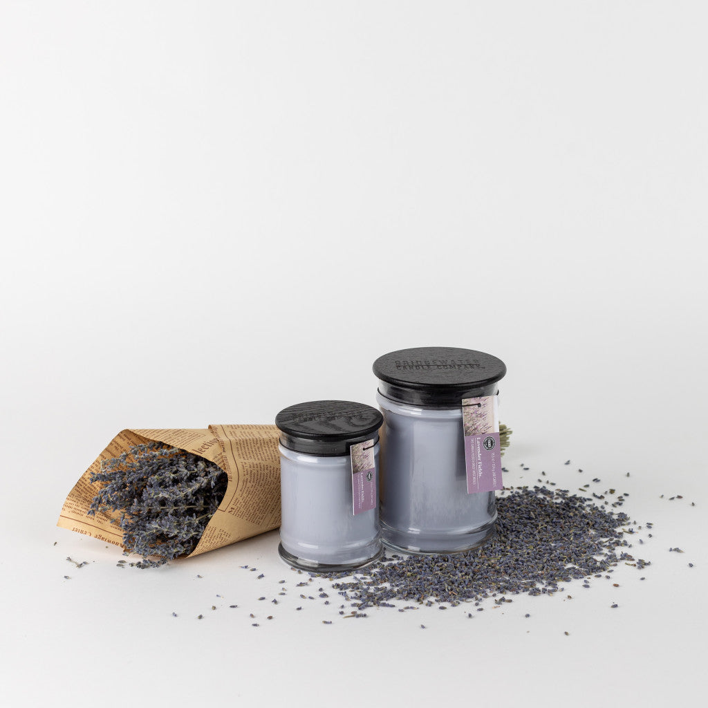 Two Bridgewater Candle Company Lavender Fields 8oz Small Jar Candles with black lids sit beside a brown paper cone of dried lavender, scattered sprigs on a white surface.