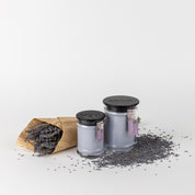Two Bridgewater Candle Company Lavender Fields 8oz Small Jar Candles with black lids sit beside a brown paper cone of dried lavender, scattered sprigs on a white surface.