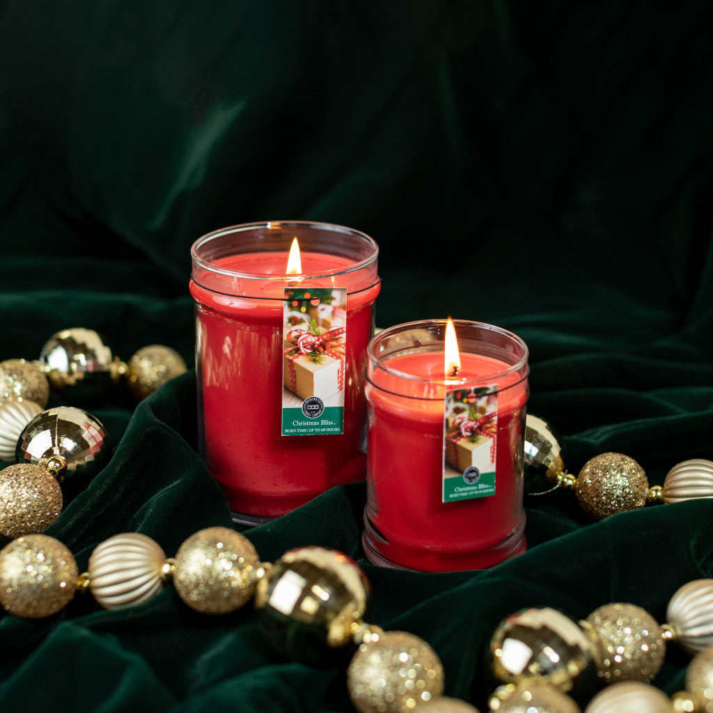 Two Christmas Bliss 8oz Holiday Jar Candles by Bridgewater Candle Company glow on dark green fabric, surrounded by gold and white Christmas ornaments to create a festive, fragrant holiday atmosphere.