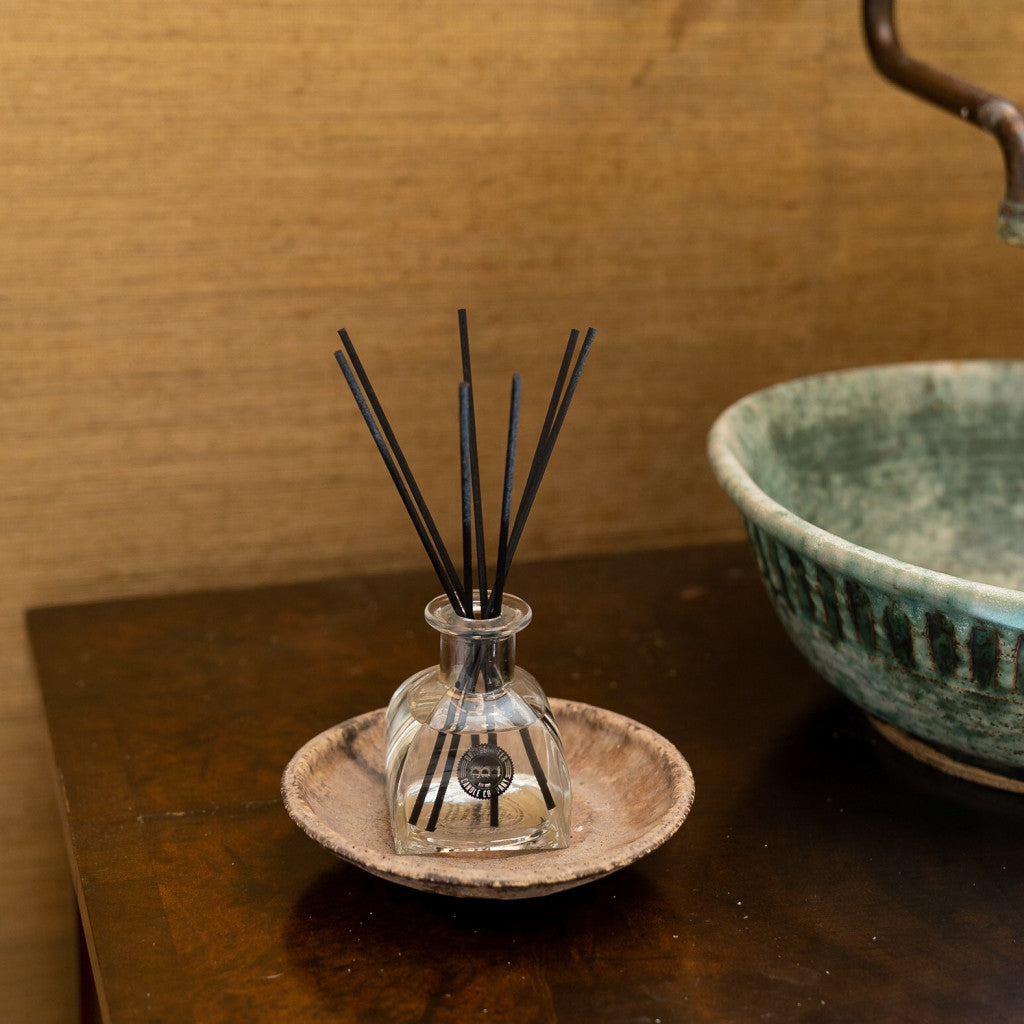 The Bridgewater Candle Company Afternoon Retreat Reed Diffuser, with black fiber reeds in a glass vessel, sits on a wooden tray beside a green ceramic bowl, filling the room with premium fragrance.