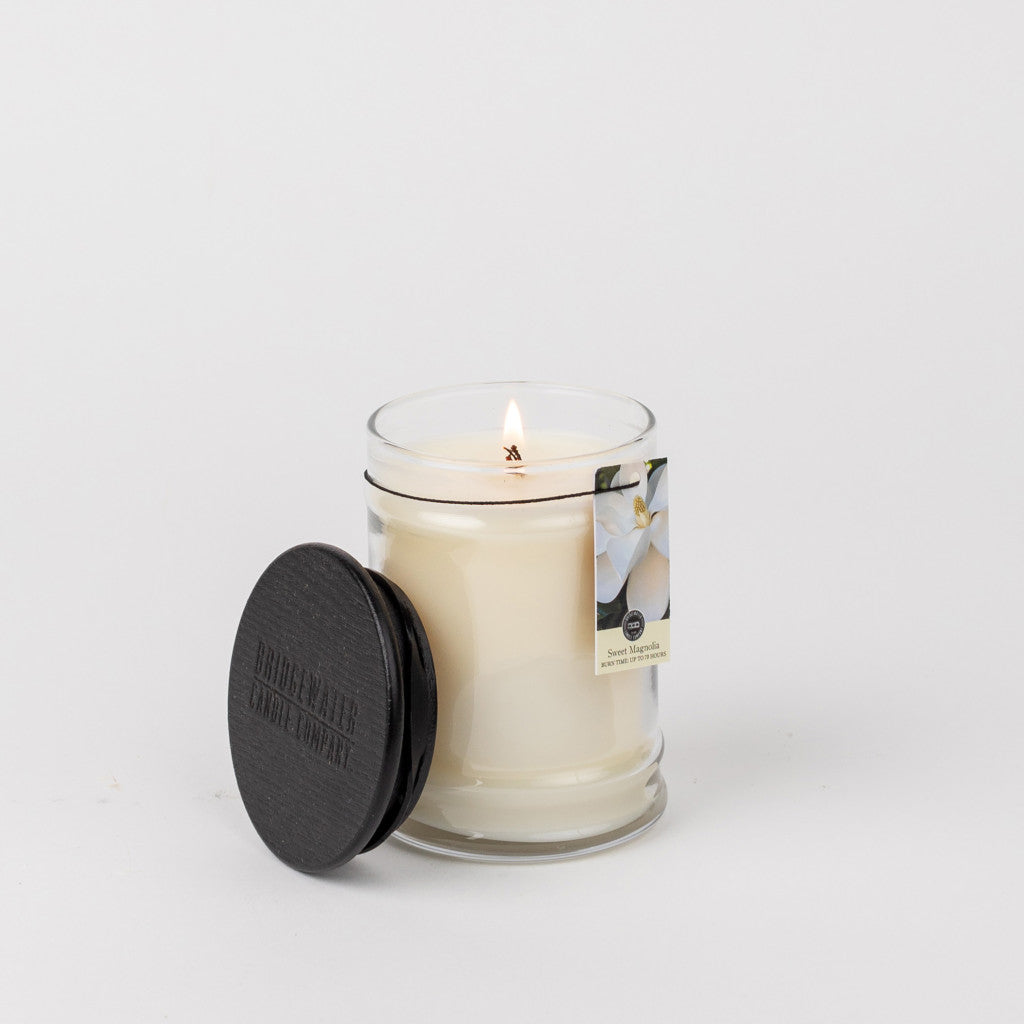 A lit Bridgewater Candle Company Sweet Magnolia 8oz jar candle sits in a clear glass jar with a black lid. The label features a white flower and “Sweet Magnolia,” suggesting a floral scent. The background is plain white.