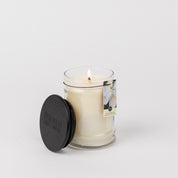 A lit Bridgewater Candle Company Sweet Magnolia 8oz jar candle sits in a clear glass jar with a black lid. The label features a white flower and “Sweet Magnolia,” suggesting a floral scent. The background is plain white.