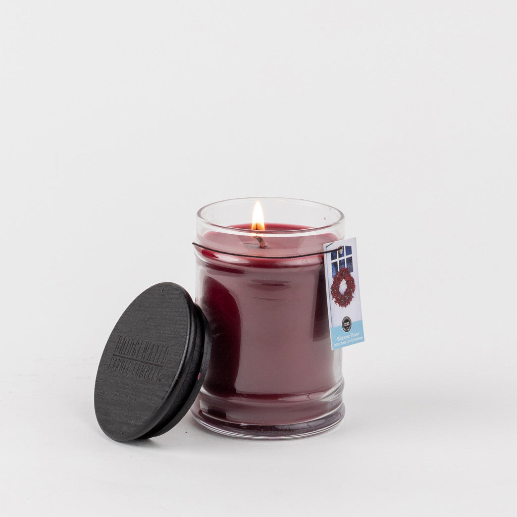 The Welcome Home 8oz Jar Candle by Bridgewater Candle Company, a small dark red soy-blend candle in a clear glass jar with a black lid and tag, is shown lit against a plain white background.