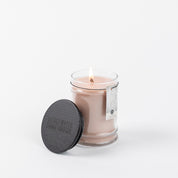 The Sweet Grace 8oz Jar Candle by Bridgewater Candle Company glows in a clear glass jar with a black textured lid beside it and a small tag attached, set against a plain white background, evoking passionate fruits and subtle patchouli.