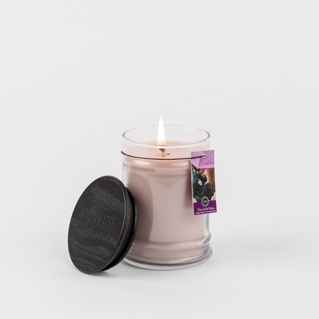 A lit, light pink Bridgewater Candle Company Kiss In The Rain 8oz Jar Candle sits on a white surface with its round black lid beside it and a purple Kiss In The Rain tag attached.