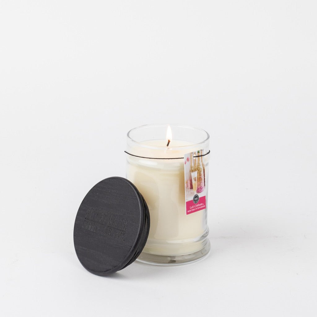 A lit Let’s Celebrate 8oz Jar Candle by Bridgewater Candle Company in a clear glass jar with a black lid sits beside it. The label displays the brand and LIGHT A CANDLE FEED A CHILD message, set against a plain white background.