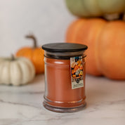The Bridgewater Candle Company Harvest Pumpkin 8oz Jar Candle, featuring orange wax and a black lid, sits on marble. Its label shows pumpkins, with blurred pumpkins behind—an ideal pick for any Pumpkin Spice Candle lover.