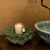A lit Comfort & Joy 8oz Small Jar Candle from Bridgewater Candle Company sits on a dark wooden surface, encircled by festive artificial pine branches. Nearby, a green ceramic bowl rests against textured tan wallpaper.