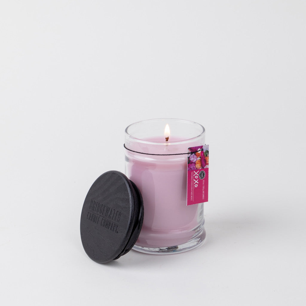 The XOXO 8oz Small Jar Candle by Bridgewater Candle Company glows in a clear glass jar with a black lid beside it. A pink floral tag is attached, all set against a plain white background.