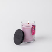 The XOXO 8oz Small Jar Candle by Bridgewater Candle Company glows in a clear glass jar with a black lid beside it. A pink floral tag is attached, all set against a plain white background.