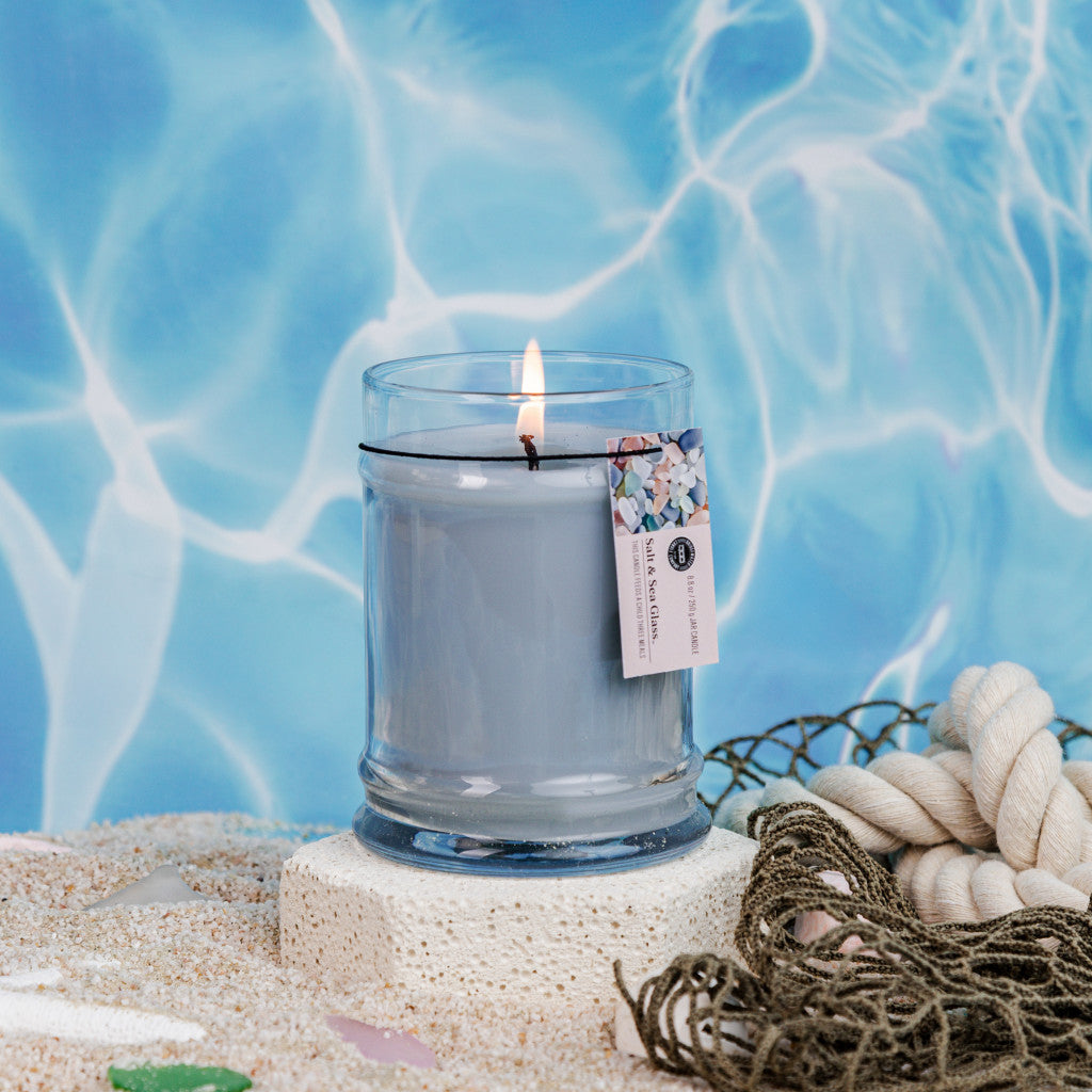The Bridgewater Candle Company Salt & Sea Glass 8oz Small Jar Candle, with soy-blend wax, sits on sand with a tag, encircled by rope, netting, and sea glass. Water reflections in the background create a serene beach-inspired scene.