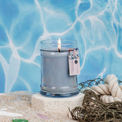 The Bridgewater Candle Company Salt & Sea Glass 8oz Small Jar Candle, with soy-blend wax, sits on sand with a tag, encircled by rope, netting, and sea glass. Water reflections in the background create a serene beach-inspired scene.
