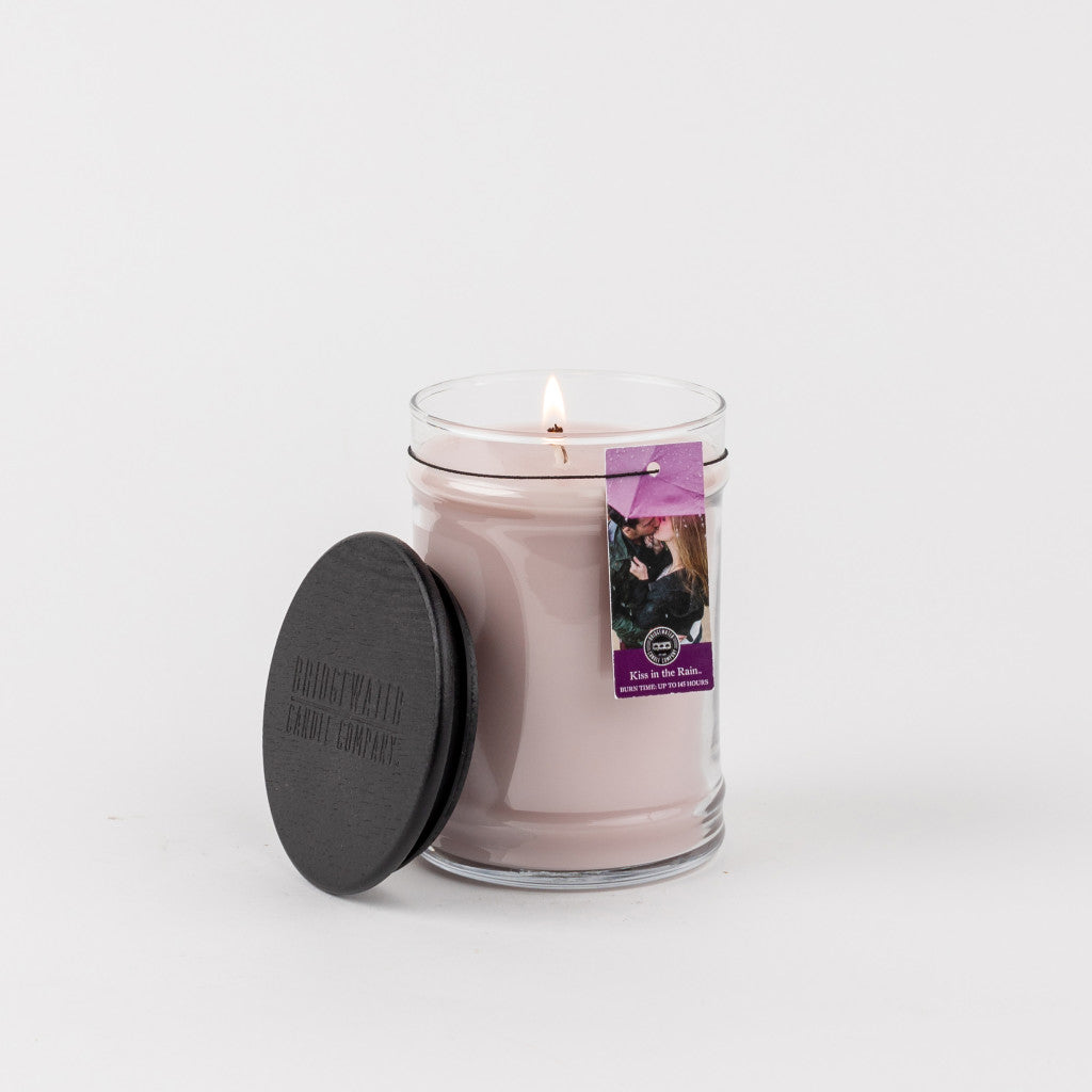 A Kiss in the Rain 18oz Large Jar Candle by Bridgewater Candle Company sits beside its black lid, featuring a purple tag with a dog and cat image. The simple white background highlights this signature fragrance.