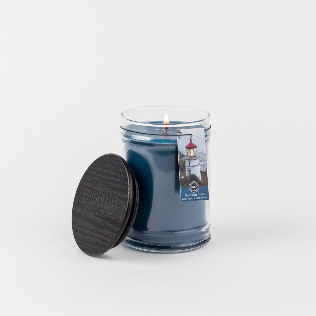 A lit Bridgewater Candle Company Nantucket Coast 18oz Large Jar Candle with a black lid sits beside it. The label shows a lighthouse, referencing the Nantucket Coast scent, all set against a plain white background.