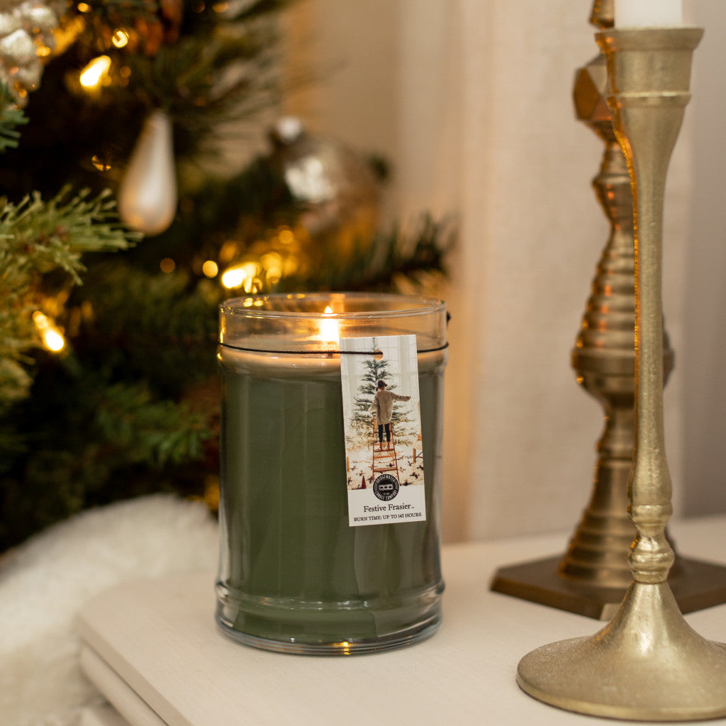A Bridgewater Candle Company Festive Frasier 18oz Large Jar Candle glows on a table beside a gold candlestick, with a sparkling Christmas tree beautifully lit and decorated in the background.