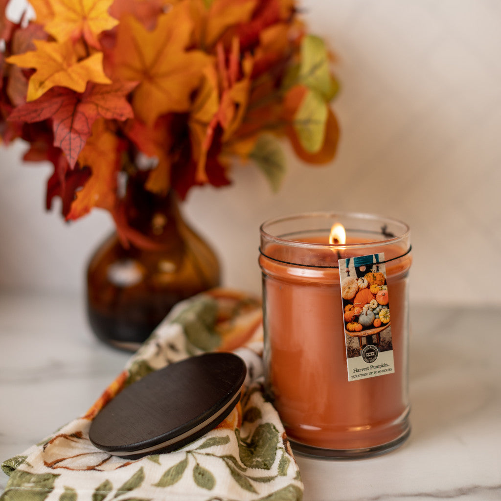 A Bridgewater Candle Company Harvest Pumpkin 18oz Large Jar Candle sits on a patterned cloth with its lid nearby. A brown vase of orange and red autumn leaves in the background adds to the cozy fall atmosphere.