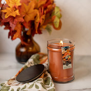 A Bridgewater Candle Company Harvest Pumpkin 18oz Large Jar Candle sits on a patterned cloth with its lid nearby. A brown vase of orange and red autumn leaves in the background adds to the cozy fall atmosphere.