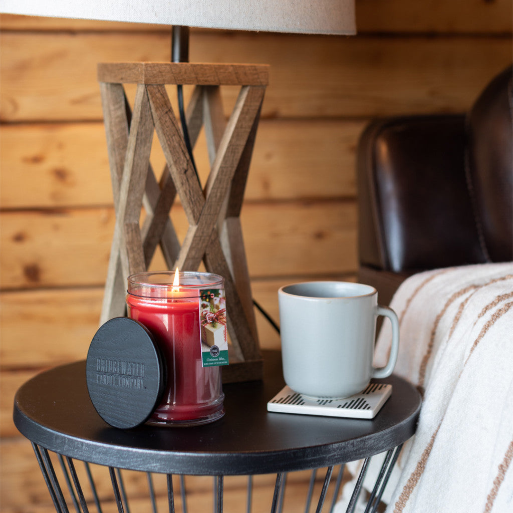 A lit Bridgewater Candle Company Christmas Bliss 18oz Large Jar, a white mug on a coaster, and a wooden lamp rest on a round black side table beside a brown leather chair and striped blanket in this cozy wooden room.