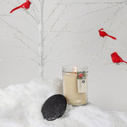 A lit Comfort & Joy 18oz Large Jar Candle by Bridgewater Candle Company sits on artificial snow with its black lid nearby, while a white tree with red cardinal ornaments in the background creates a festive ambiance.