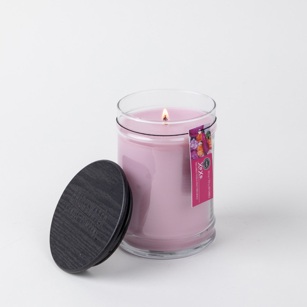 A lit Bridgewater Candle Company XOXO 18oz Large Jar Candle in a clear glass jar with a black wooden lid to the side. The soy-blend candle has a floral label with text and is set against a plain white background.