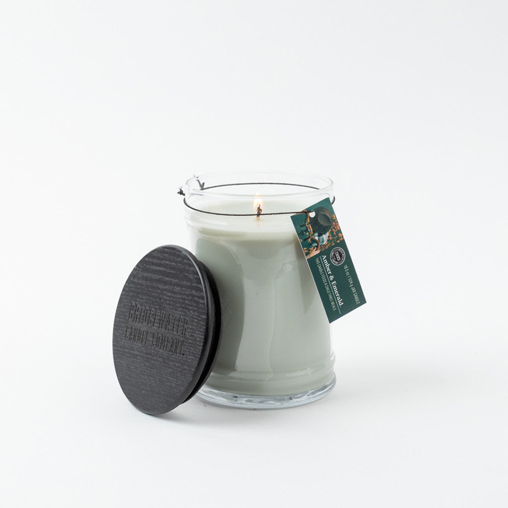 The Bridgewater Candle Company Amber & Emerald 18oz Large Jar Candle, made with soy-blend wax, sits in a clear glass jar with a black wooden lid and green product tag, all displayed against a white background.