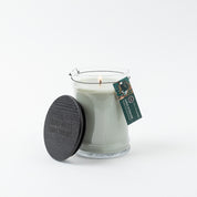 The Bridgewater Candle Company Amber & Emerald 18oz Large Jar Candle, made with soy-blend wax, sits in a clear glass jar with a black wooden lid and green product tag, all displayed against a white background.