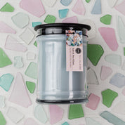 The Bridgewater Candle Company Salt & Sea Glass 18 oz Large Jar Candle features a light blue soy-blend wax in a glass jar with black lid, set against fabric with pastel green, pink, and blue sea glass shapes.