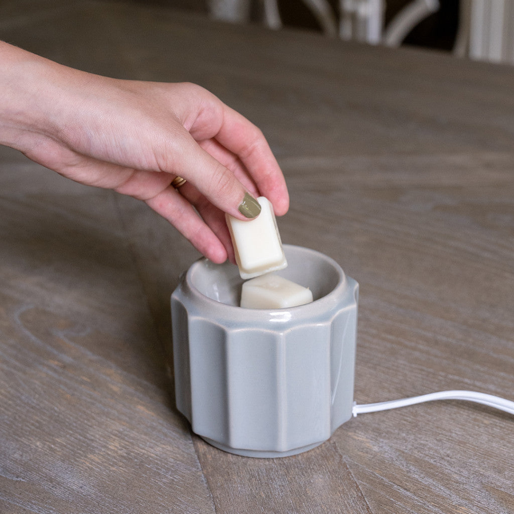 A hand places a white wax melt into the Bridgewater Candle Company Wax Warmer-Grey Banded on a wooden table. This electric warmer, with a white cord, is ideal for using wax melts and fragrance oils in any room.