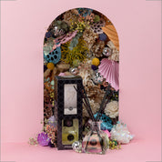 The Bridgewater Candle Company Sweet Grace Reed Diffuser, featuring black reeds and its box, is showcased before a vibrant floral backdrop on a pastel pink surface—bringing elegant fragrance to your home.