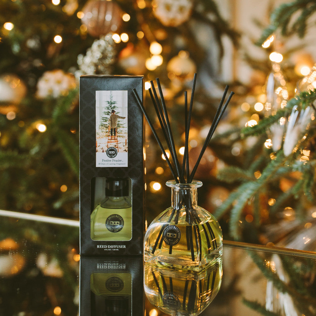A Bridgewater Candle Company Festive Frasier Reed Diffuser with fiber reeds stands next to its box on a reflective surface, framed by a glowing Christmas tree and warm lights that create a cozy, festive holiday mood.