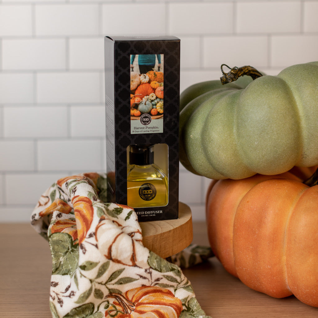 A Bridgewater Candle Company Harvest Pumpkin Reed Diffuser sits on a wooden surface beside two decorative pumpkins and a pumpkin-patterned cloth, bringing cozy fall fragrance against a white subway tile backdrop.