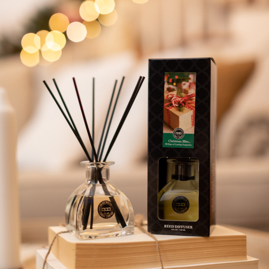 A Bridgewater Candle Company Christmas Bliss Reed Diffuser with black reeds sits on wood by its box, surrounded by soft holiday lights and cozy décor for a festive touch.