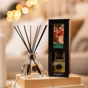 A Bridgewater Candle Company Christmas Bliss Reed Diffuser with black reeds sits on wood by its box, surrounded by soft holiday lights and cozy décor for a festive touch.