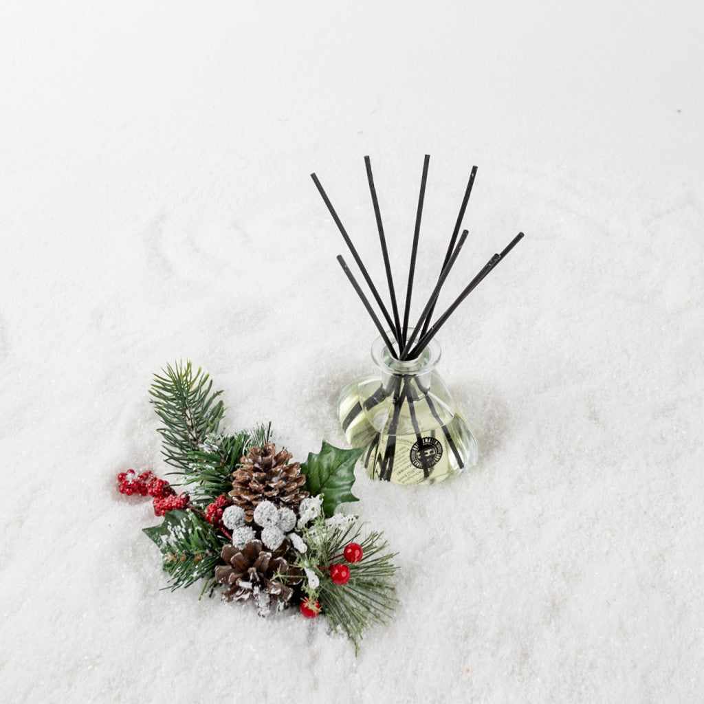 A Comfort & Joy Reed Diffuser by Bridgewater Candle Company with sleek black reeds sits on snowy pinecones, red berries, and snow-dusted pine branches, adding inviting fragrance to the festive scene.