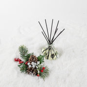 A Comfort & Joy Reed Diffuser by Bridgewater Candle Company with sleek black reeds sits on snowy pinecones, red berries, and snow-dusted pine branches, adding inviting fragrance to the festive scene.